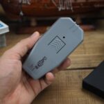 Ultradźwiękowy odstraszacz psów Dazer II w dłoni mężczyzny. Widoczny przycisk wyzwalania i logo. W tle stół z rozmaitymi przedmiotami.