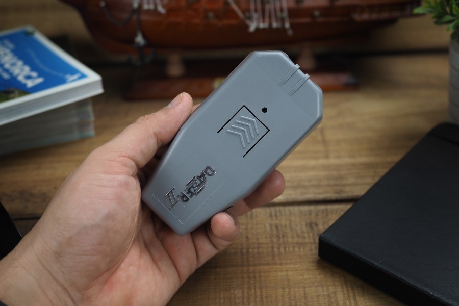 Ultradźwiękowy odstraszacz psów Dazer II w dłoni mężczyzny. Widoczny przycisk wyzwalania i logo. W tle stół z rozmaitymi przedmiotami.