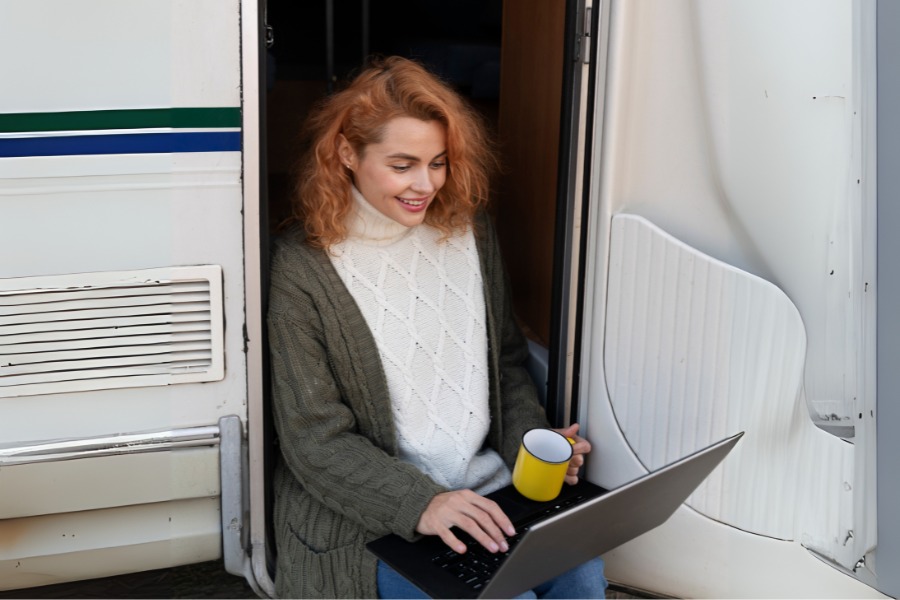 Uśmiechnięta kobieta siedzi w drzwiach kampera z laptopem na kolanach i kubkiem w dłoni, korzystając z Internetu podczas podróży.