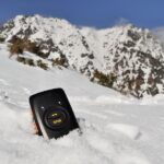 Czarny nadajnik GPS z przyciskiem SOS zakopany do połowy w śniegu, w tle wysokie góry.