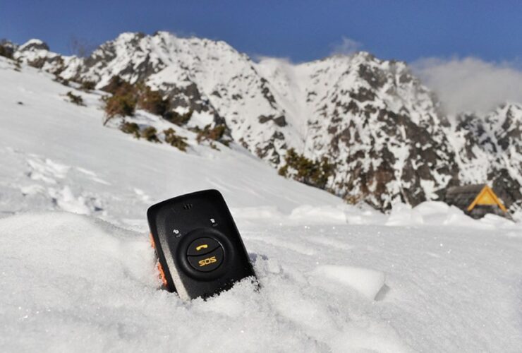 Czarny nadajnik GPS z przyciskiem SOS zakopany do połowy w śniegu, w tle wysokie góry.