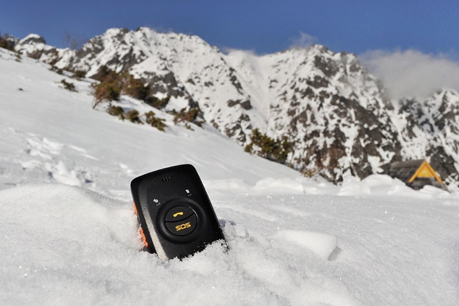 Czarny nadajnik GPS z przyciskiem SOS zakopany do połowy w śniegu, w tle wysokie góry.
