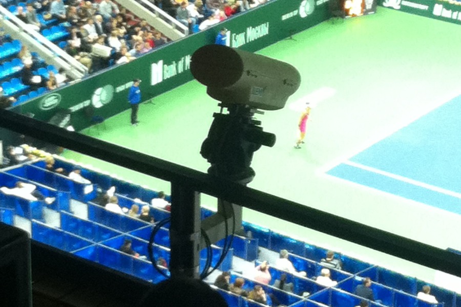 Jedna z kamer, która obsługuje system Hawk-Eye, przymocowana do barierki na górnej części areny. W tle widoczny kort tenisowy, kibice i mecz.