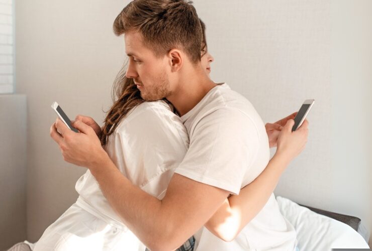 Mężczyzna i kobieta przytulają się na łóżku, każde z nich używa swojego telefonu. Pokazuje to, że tak naprawdę są oni sobie obojętni.