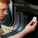 Mężczyzna trzyma w ręku biały lokalizator GPS. Nachyla się w samochodzie i montuje nadajnik pod kierownicą w kokpicie.