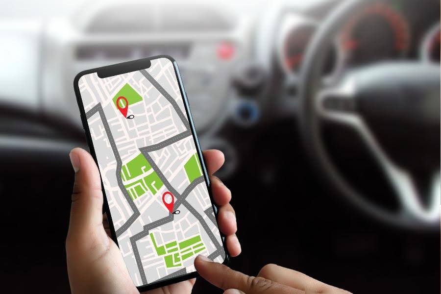 Osoba trzymająca smartfona z otwartą aplikacją map, pokazującą trasę i dwa znaczniki lokalizacji, w tle wnętrze samochodu.