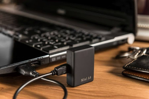 Podsłuch Mini A8 podłączony do laptopa za pomocą kabla USB, leżący na drewnianym biurku obok portfela i kluczy.