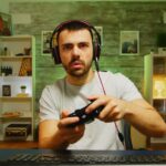 Zaskoczony mężczyzna w słuchawkach trzyma pada do konsoli – wygląda, jakby nie dowierzał temu, co widzi na ekranie.