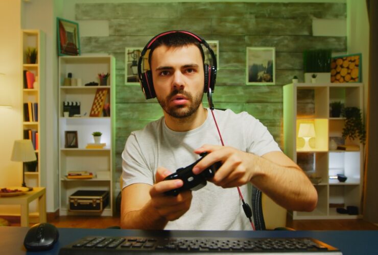 Zaskoczony mężczyzna w słuchawkach trzyma pada do konsoli – wygląda, jakby nie dowierzał temu, co widzi na ekranie.