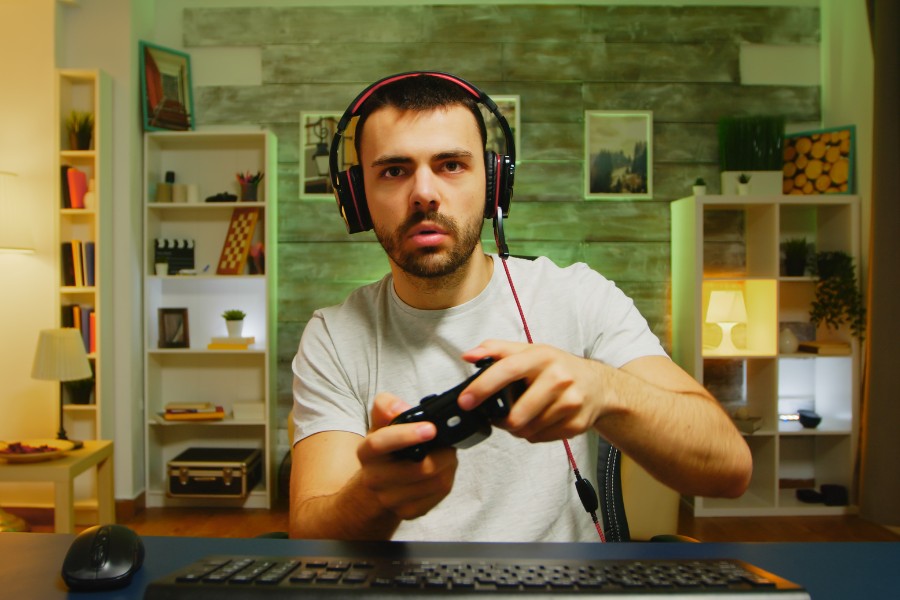 Zaskoczony mężczyzna w słuchawkach trzyma pada do konsoli – wygląda, jakby nie dowierzał temu, co widzi na ekranie.