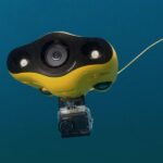 Na zdjęciu został przedstawiony żółty dron podwodny z kamerą zanurzony w wodzie, podłączony przewodem.