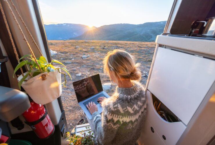 Kobieta siedząca w kamperze i pracująca na laptopie, w otoczeniu kamienistego krajobrazu.