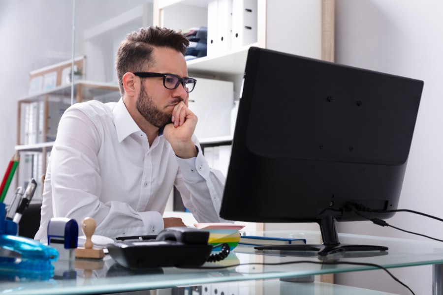 Zamyślony mężczyzna w koszuli i okularach podpiera się ręką. Siedzi w biurze i patrzy się w monitor.