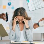 Zestresowana kobieta przy biurku, otoczona przez współpracowników pokazujących dokumenty i telefon.