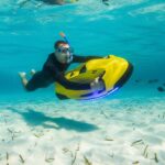 Nurek w skafandrze, goglach i z rurką do snorkelingu w ustach znajduje się pod wodą i trzyma żółty skuter podwodny. Wokół błękit wody, piaszczyste dno i ryby.