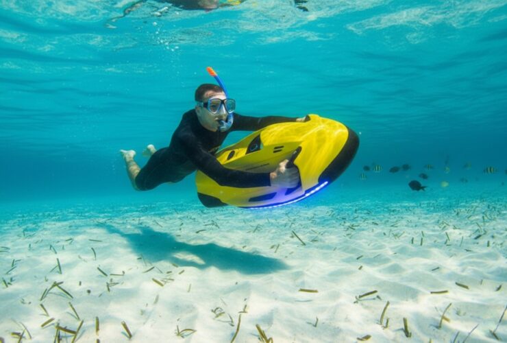 Nurek w skafandrze, goglach i z rurką do snorkelingu w ustach znajduje się pod wodą i trzyma żółty skuter podwodny. Wokół błękit wody, piaszczyste dno i ryby.