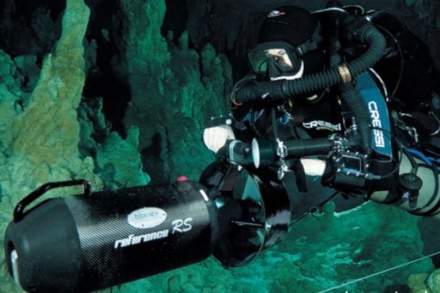 Nurek w profesjonalnym skafandrze i masce eksploruje teren pod wodą. Obiema rękami trzyma profesjonalny skuter wodny.