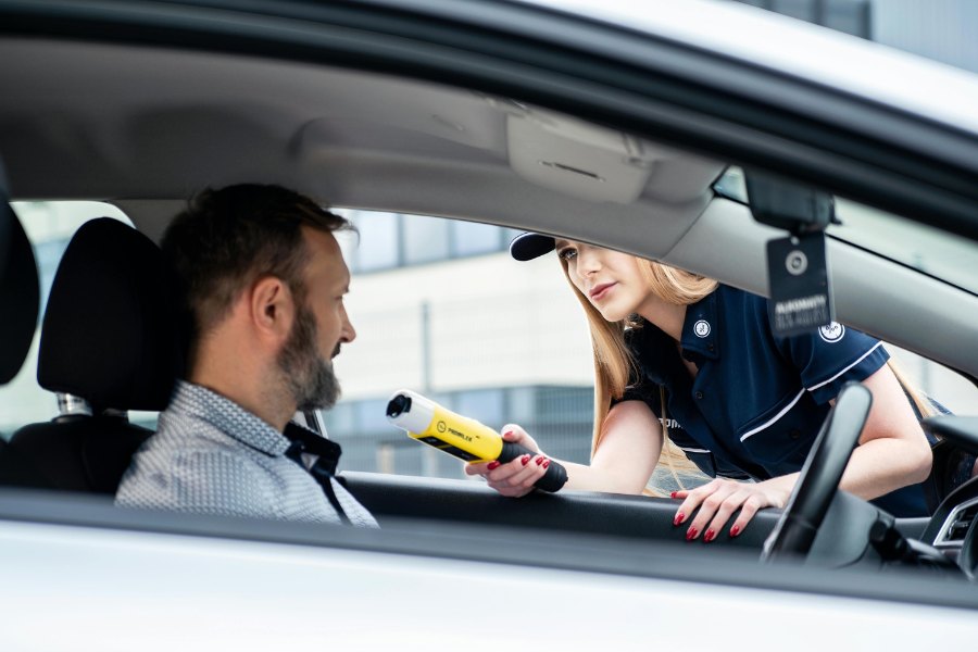 Na grafice został przedstawiony proces pomiaru profesjonalnym alkomatem przez służby mundurowe.