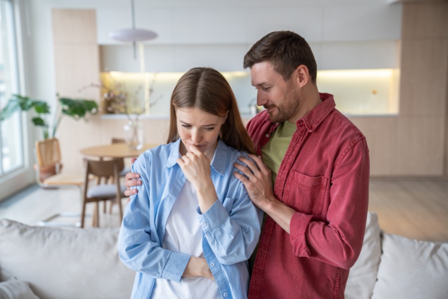 Mężczyzna w czerwonej koszuli obejmuje swoją partnerkę, pocieszając ją. Kobieta w niebieskiej koszuli płacze i podpiera głowę. W tle jasny pokój dzienny.