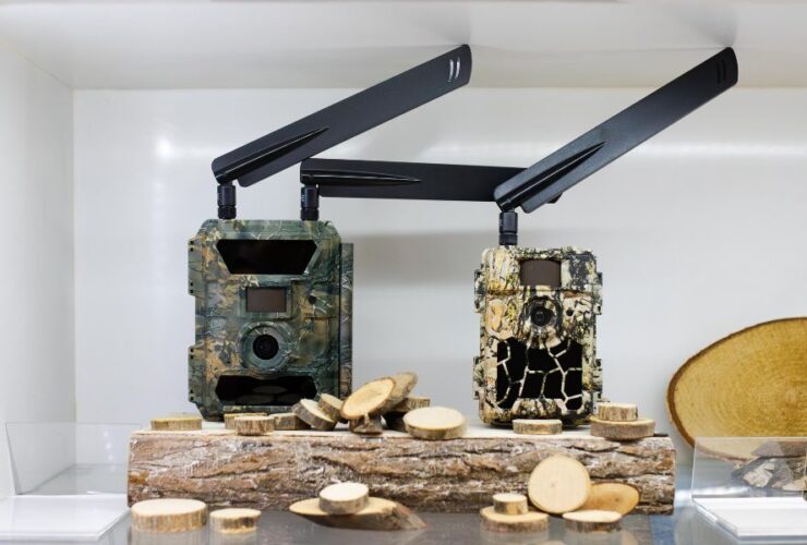 Dwie fotopułapki z antenami ustawione na drewnianych klockach, obudowy w kamuflażu, widoczne obiektywy i czujniki ruchu