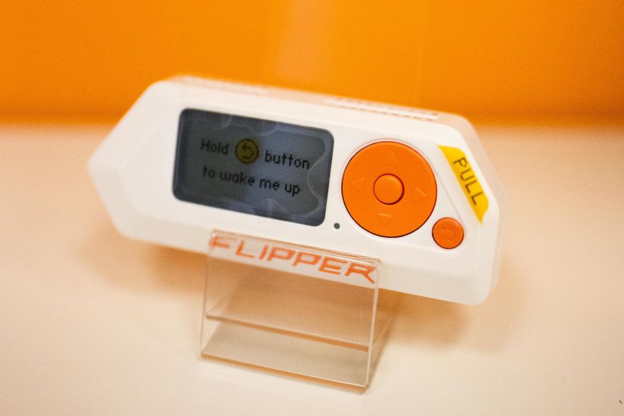 Flipper Zero z pomarańczowymi przyciskami stoi na przezroczystej podstawce na tle pomarańczowego tła.