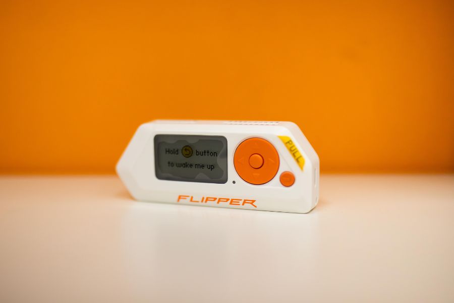 Biały Flipper Zero z pomarańczowymi przyciskami leży na jasnej powierzchni na tle intensywnie pomarańczowego tła.