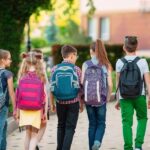 Grupa sześciu uczniów z kolorowymi plecakami idzie chodnikiem w kierunku budynku szkoły w słoneczny dzień.