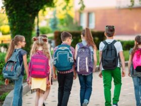 Grupa sześciu uczniów z kolorowymi plecakami idzie chodnikiem w kierunku budynku szkoły w słoneczny dzień.