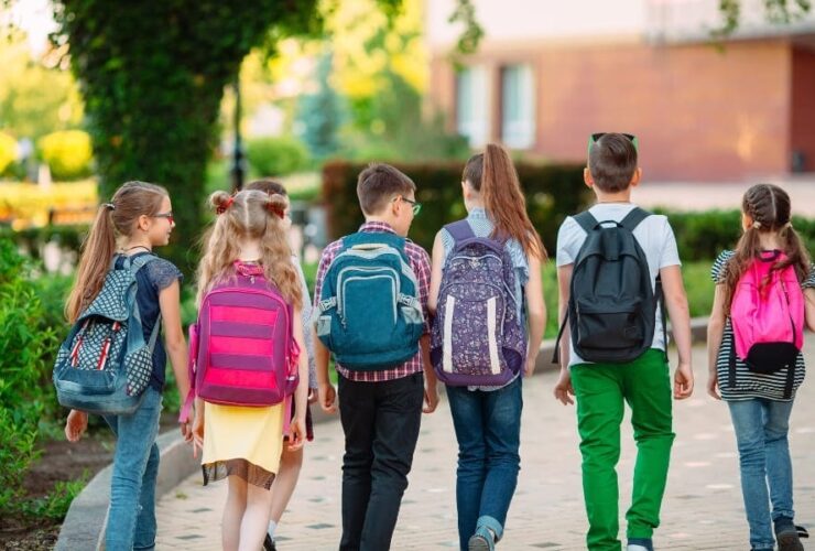 Grupa sześciu uczniów z kolorowymi plecakami idzie chodnikiem w kierunku budynku szkoły w słoneczny dzień.