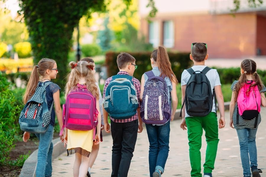 Grupa sześciu uczniów z kolorowymi plecakami idzie chodnikiem w kierunku budynku szkoły w słoneczny dzień.